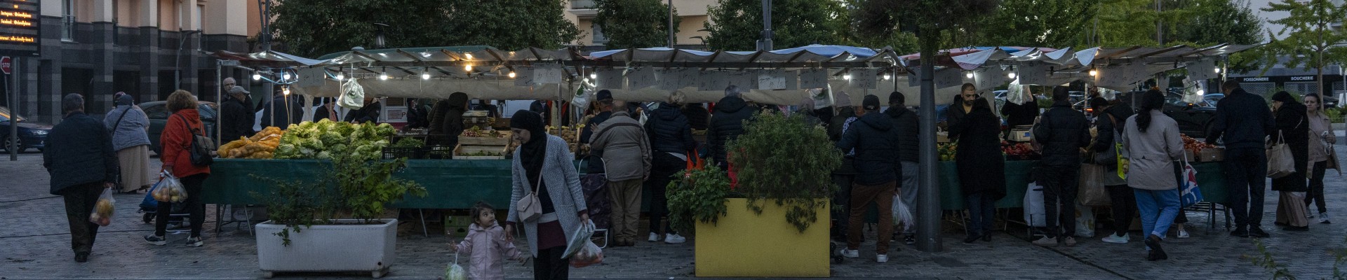 Des stands de marché en fond, quelques habitants marchant sur l'esplanade devant dont une femme tenant un enfant par la main