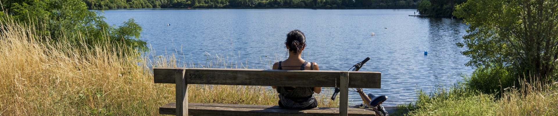 Une femme de dos, assise sur un banc, son vélo à côté d'elle, regarde le lac