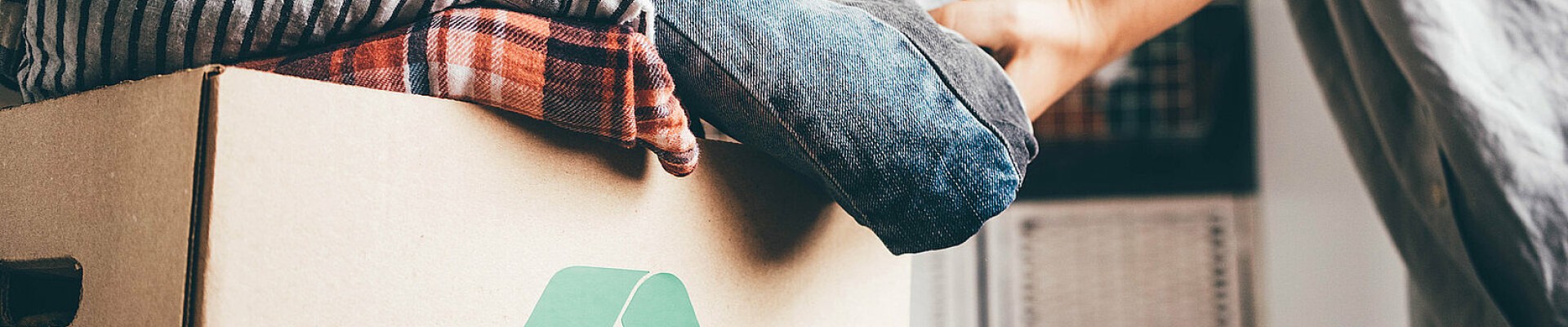 Des vêtements dans un carton avec le logo recyclage, et une personne dont ne voit que les bras qui les inspectent