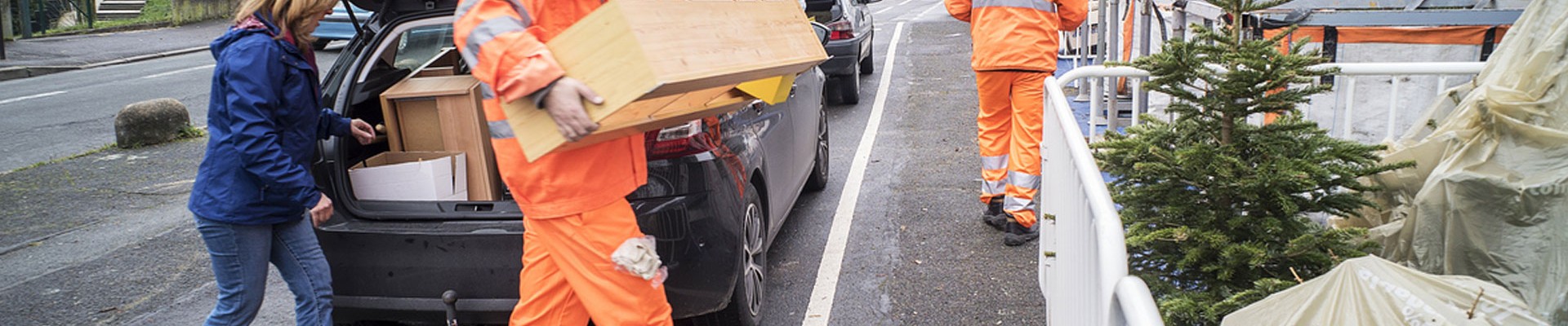 Agents de déchèterie mobile aidant un particulier à déposer des morceaux de meubles dans des enclos prévus pour ce type de déchet.