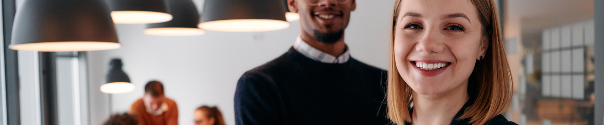 2 personnes souriantes 1 femme et 1 homme dans une salle de réunion, d'autres personnes en fond