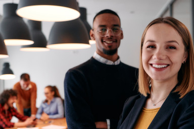 2 personnes souriantes 1 femme et 1 homme dans une salle de réunion, d'autres personnes en fond