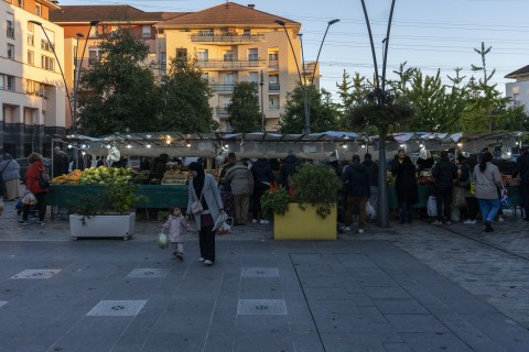 Des stands de marché en fond, quelques habitants marchant sur l'esplanade devant dont une femme tenant un enfant par la main