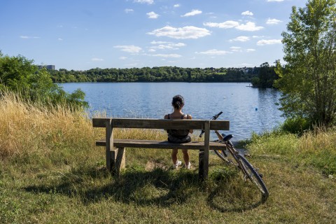 Une femme de dos, assise sur un banc, son vélo à côté d'elle, regarde le lac
