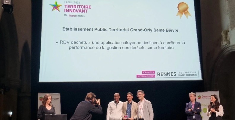 Des personnes souriantes sur un podium surmonté d'une projection indiquant le label Territoire innovant 2025 des Interconnectés et indiquant que le Grand-Orly Seine Bièvre est lauréat pour "RDV déchets : une application citoyenne destinée à améliorer la performance de la gestion des déchets sur le territoire" - Agrandir l'image, fenêtre modale